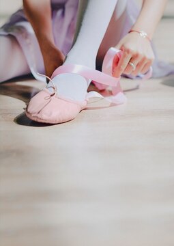Ballet dancer tying pink pointe shoes on a wooden floor, close-up.