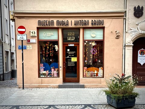 A front-view photograph of the entrance to the Museum of Soap and History of Dirt in Bydgoszcz, displaying its main sign, wooden glass door, decorated display windows, street sign, and a flower  