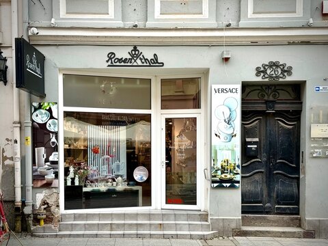 A front-view photograph of a Rosenthal store facade with big display windows, white frames, black brand logos, a Versace advertising panel, and an ornate wooden door next to gray stone steps.