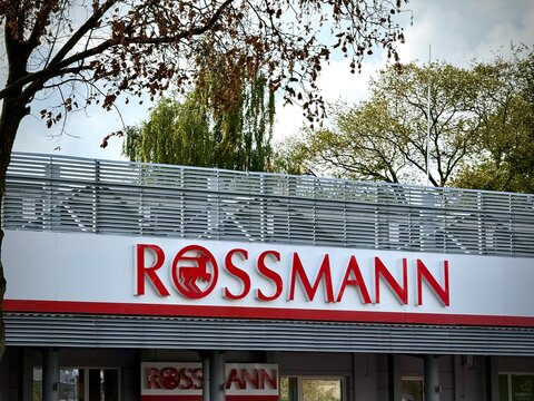 A horizontal view of the Rossmann drugstore signage featuring red lettering and horse logo, mounted on a building facade with metal louvers under a bright sky with trees in the background.