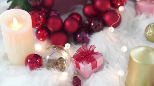 various decorative elements arranged on a white faux-fur base, ranging from softly glowing electric candles and a pink gift box with a ribbon to a wreath adorned with shiny red Christmas baubles.