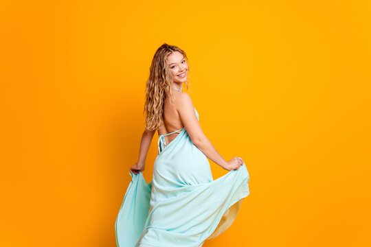 Smiling woman in a flowing light blue backless dress twirling against a vibrant orange background conveying summer joy, movement, youth, playful fashion and confident elegance