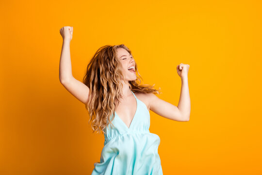 Young woman in blue dress celebrating with raised arms and joyful smile on bright yellow background, expressing excitement, confidence and carefree summer fun