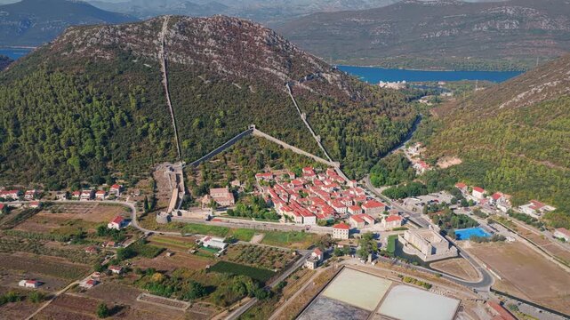 Cinematic aerial footage of Ston village with ancient fortress walls, vibrant rooftops, and geometrically patterned salt pans under clear skies, surrounded by lush hills and blue water.