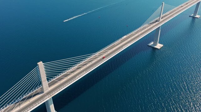 Drone shot of Pelje&scaron;ac Bridge spanning crystal blue waters, with boats gliding below. Bright daylight highlights modern architecture and sweeping coastal scenery from above.