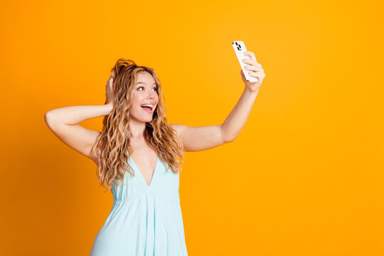 Happy young woman taking a selfie with smartphone on bright yellow background wearing a light summer dress and smiling portrait expressing joy and confidence