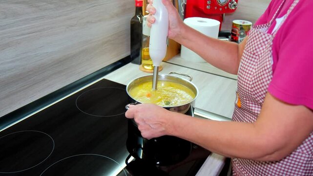 Hands using electric hand blender to puree soup in stainless steel pot on induction cooktop creating smooth texture and comforting home cooking scene.