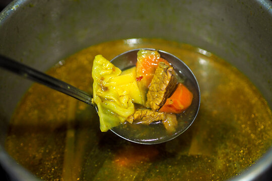 metal ladle scooping hot beef and vegetable soup with broth inside a large cooking pot