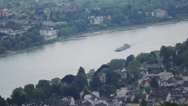 A large cargo boat is traveling down the Rhine river near a city of Bonn, aerial shot