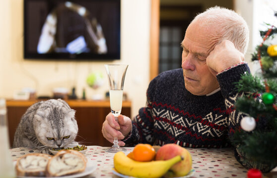 Lonely elderly man feeding cat at table during celebration of New Year