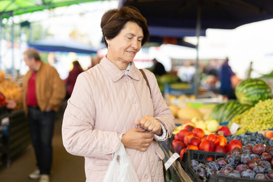 Smiling elderly female customer choosing organic plums while walking at local bazaar