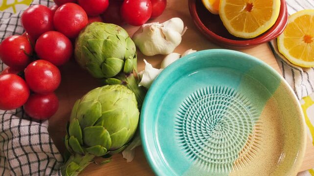 Fresh artichokes, ripe tomatoes, garlic and sliced lemon arranged around a traditional Spanish ceramic grater bowl on a kitchen table. Healthy Mediterranean ingredients prepared for homemade cooking