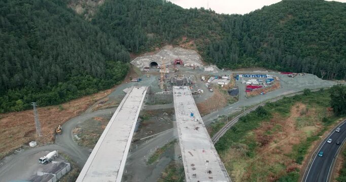 A construction site with a bridge and tunnel digging in the background. The bridge is being built over a river. The construction site is surrounded by trees and the sky is blue
