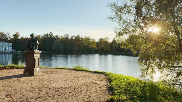 A peaceful monument stands overlooking a serene lake. A smooth camera glides past trees in Catherine Park, Pushkin, Saint Petersburg, evoking calm and wonder.