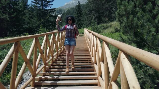 A woman wearing a plaid shirt and blue jeans is walking across a wooden stairs bridge. She is carrying a backpack and hat