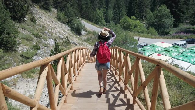 A woman wearing a plaid shirt and blue jeans is walking across a wooden stairs bridge. She is carrying a backpack and hat