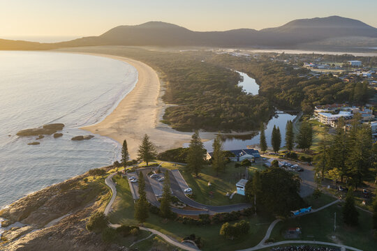 Aerial view of Front Beach and Saltwater Creek beside a coastal town, with rocky shoreline, grassy parkland and winding walking paths in the foreground. 