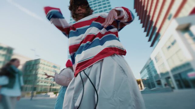 A woman in a striped shirt gestures toward the camera while dancing with a multiracial hip hop dance group in a modern outdoor plaza in Barcelona, Spain.