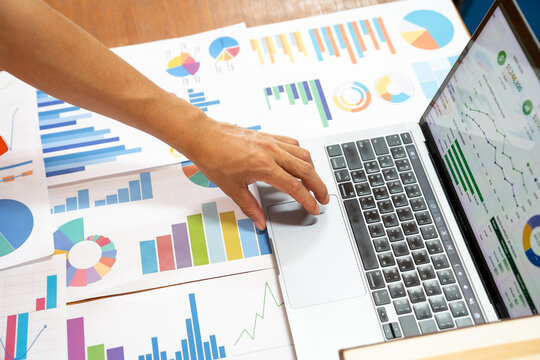 Top down view of a person using a laptop computer surrounded by numerous printed sales reports and colorful financial charts