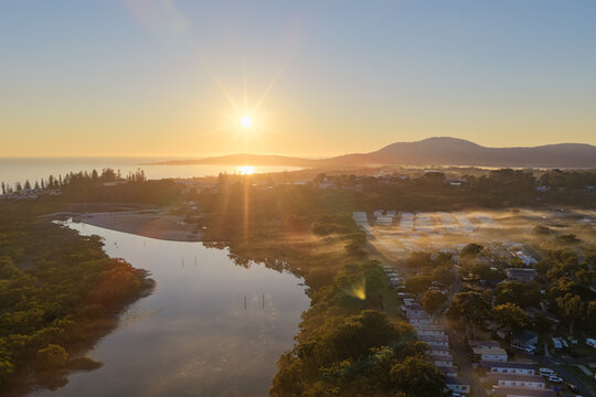 Aerial view of Back Creek bordered by trees and a caravan park, with morning mist drifting between the cabins and vegetation. A sunstar rises above the ocean, casting golden light across the landscape