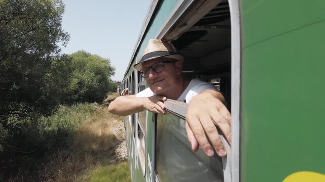 Traveler man with hat and glasses is sitting in a train window, pointing out the window. The train is on a track next to a field