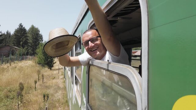 A man is waving from the window of a train open window traveling on track. The train is green and is traveling down the tracks