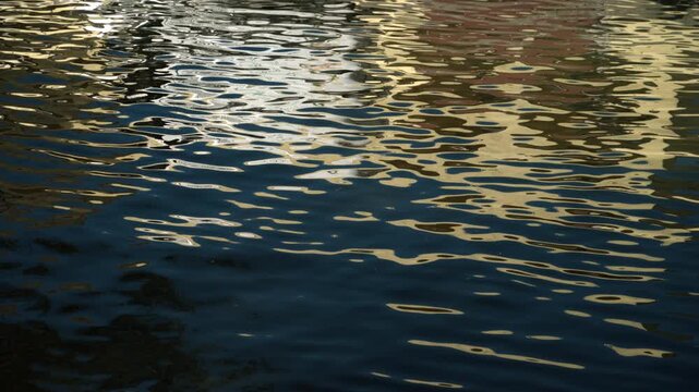 Abstract water reflection of colorful city buildings distorted by gentle river ripples. Warm golden sunlight and deep blue shadows create a painterly fluid texture. 4K.
