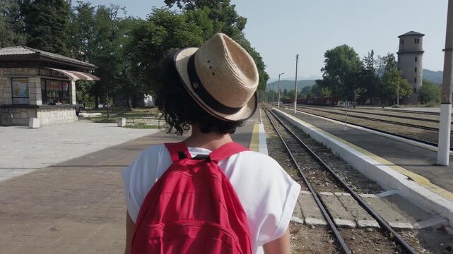 A woman wearing a straw hat and a red backpack is standing on a train platform. She is smiling and looking at the camera