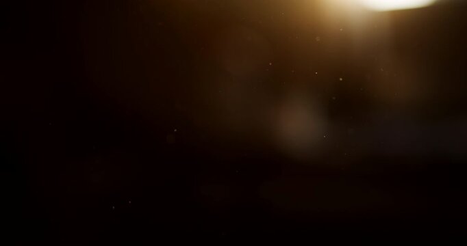 Golden light flare glowing from the top edge of a dark background, with tiny dust particles floating in the air and soft warm bokeh, abstract overlay with copy space, 4K