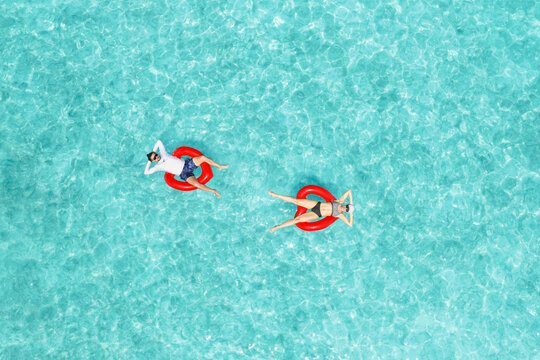 Aerial view of a man and a woman floating on red heart-shaped rings in the crystal clear turquoise water of Vashafaru, Haa Alif Atoll, Maldives.
