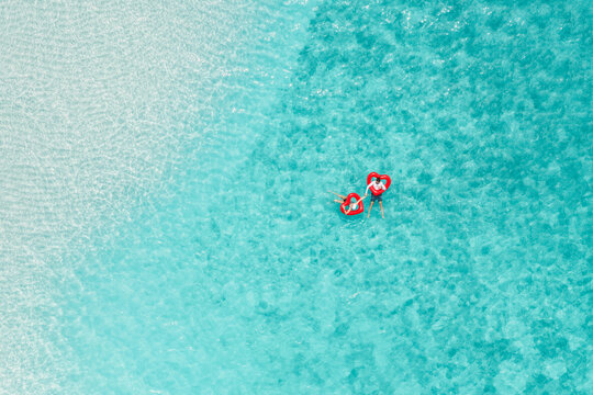 Aerial view of a couple relaxing on red heart-shaped floats in the clear turquoise water of the Indian Ocean in Vashafaru, Haa Alif Atoll, Maldives.