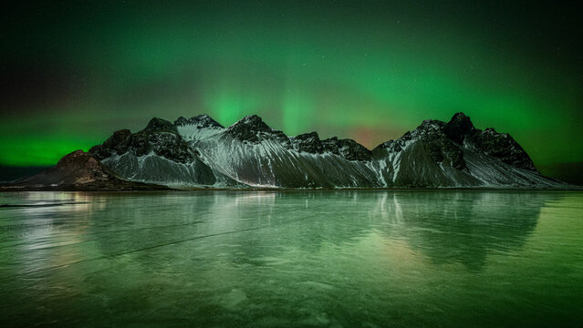 Vestrahorn mountain and green Northern Lights shimmering across a frozen inlet in winter in Iceland