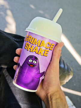 Hand holding a purple McDonald's Grimace Shake cup with white lid and straw, yellow lettering and Grimace character artwork, outdoors on sunlit asphalt pavement, closeup