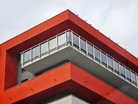 Modern red apartment building corner with glass balcony railings, grey concrete floors and overcast sky, low angle upward view of geometric facade architecture, minimal urban design