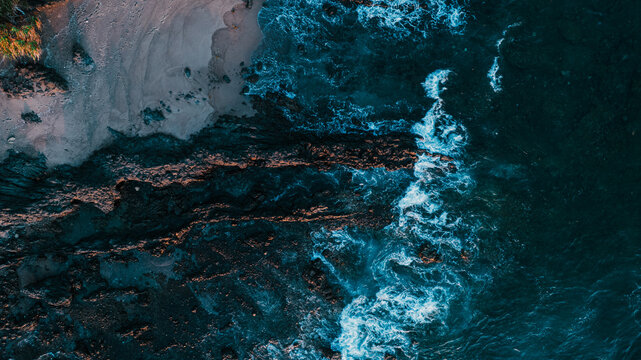 Aerial view of dark volcanic rocks meeting the turquoise ocean waves along a sandy beach in Bajawa, Nusa Tenggara Timur, Indonesia.