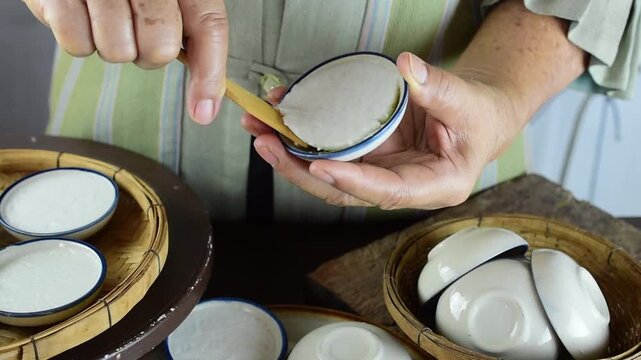 Close-up of senior woman using flat stick to scrape warm traditional Thai steamed coconut pudding from small ceramic cup in local kitchen.