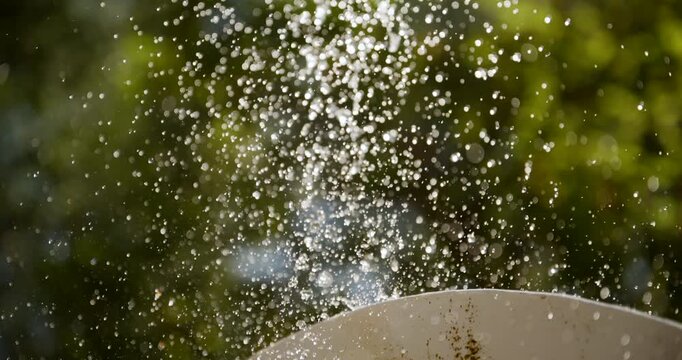 Close-up of water droplets splashing and suspended mid-air above a stone fountain basin with lush green bokeh background. 4K slow motion.