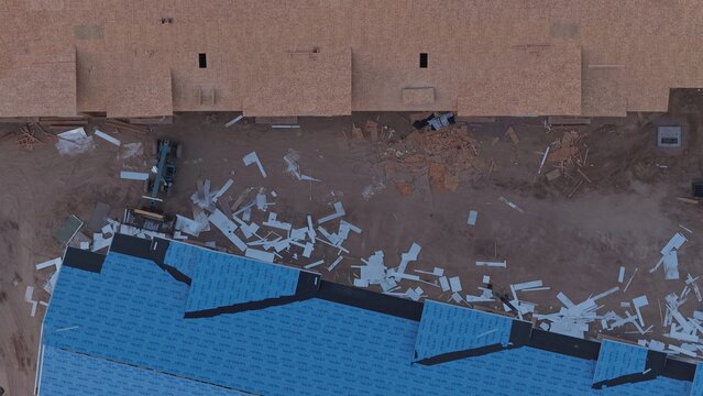 Aerial view of a construction site with blue roof underlayment, wooden framing, and scattered debris on dirt ground in Greeley, Colorado, United States.