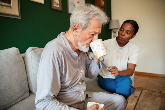 Caregiver helping senior man sip from a mug at home