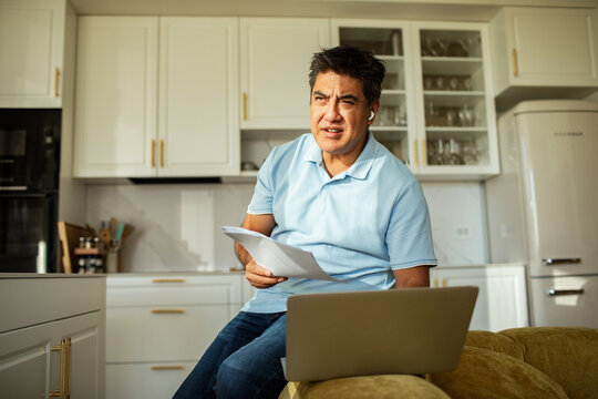 Concerned man with earbuds reviewing paperwork on laptop in kitchen