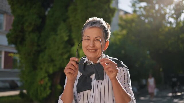 A smiling mature woman stands outdoors in soft afternoon light wearing a striped shirt and sweater over her shoulders. The candid portrait conveys warmth, confidence, relaxed urban lifestyle and posit