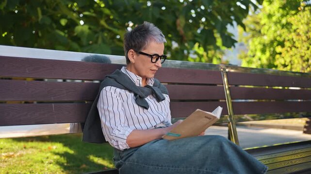Elderly woman with short gray hair reads a book on a wooden bench in a park, with lush green trees and sunlight creating a serene atmosphere
