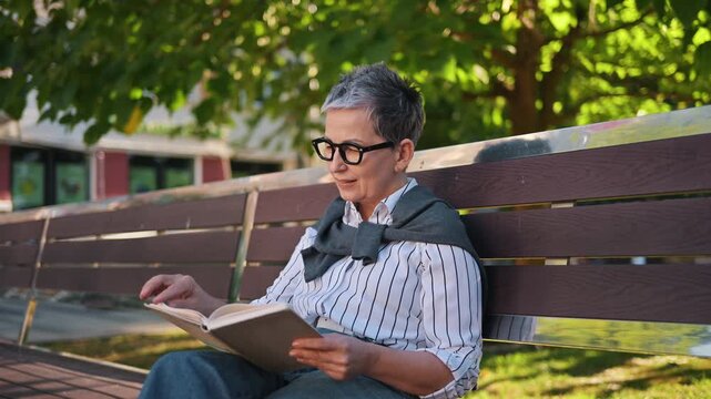 Senior woman with gray hair and glasses enjoys reading a book on a wooden bench in a park, with greenery and sunlight creating a serene atmosphere