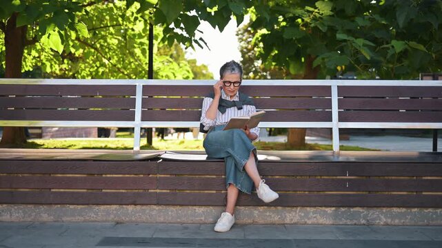 Senior woman with gray hair and glasses enjoys reading a book on a wooden bench in a park, with greenery and sunlight creating a serene atmosphere