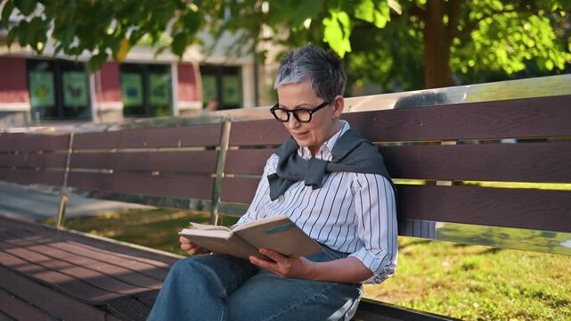 Senior woman with gray hair and glasses enjoys reading a book on a wooden bench in a park, with greenery and sunlight creating a serene atmosphere
