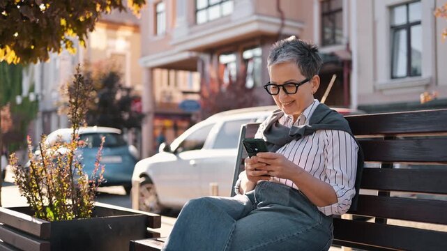 Mature woman with short gray hair wearing glasses and striped shirt sits on a bench outdoors, using a smartphone with parked cars and greenery in the background