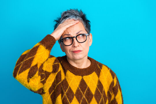 Mature woman in glasses wearing yellow argyle sweater looking puzzled and holding her head with a thoughtful, frustrated expression against a bright blue background