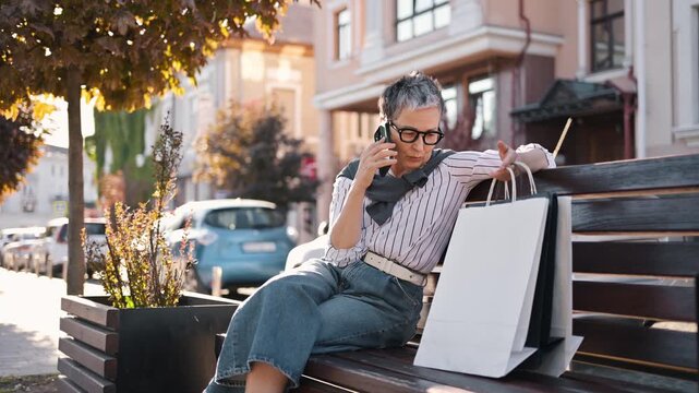Senior woman with gray hair and glasses engages in a phone conversation while seated on a bench in a sunny urban park with cars visible in the background