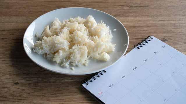 White cooked rice served on a ceramic plate with a calendar beside it, representing meal planning, diet scheduling, nutrition, and daily food consumption.