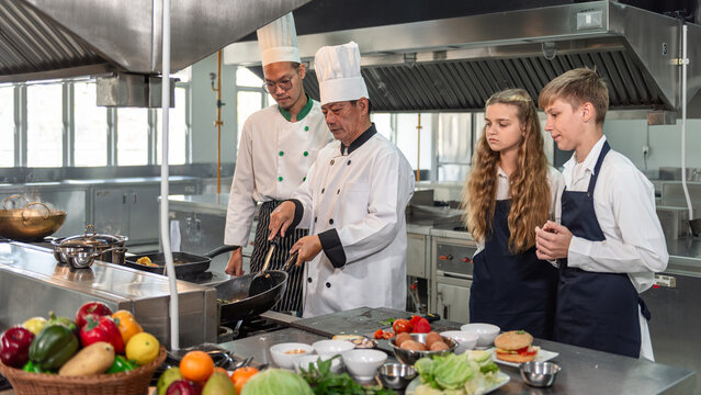 Professional Chef Teaching Culinary Students in a Commercial Kitchen, Demonstrating Wok Cooking Technique While Junior Chefs Observe a Practical Hands On Chef School Lesson